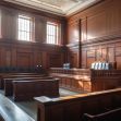 A view of an empty courtroom, showcasing wooden benches and the judge's bench illuminated by natural light. A view of an empty courtroom, showcasing wooden benches and the judge's bench illuminated by natural light.