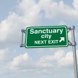 A green road sign indicating "Sanctuary City" with an arrow pointing to the right, against a sky background with clouds. A green road sign indicating "Sanctuary City" with an arrow pointing to the right, against a sky background with clouds.