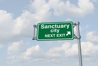 A green road sign indicating "Sanctuary City" with an arrow pointing to the right, against a sky background with clouds.
