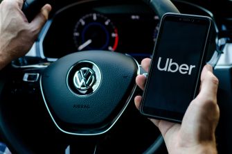 A close-up of hands holding a smartphone displaying the Uber logo while gripping a Volkswagen steering wheel.