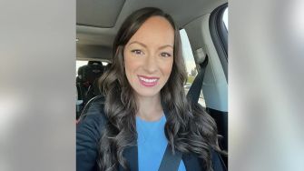 A woman smiling in a car, wearing a blue top and a black blazer.