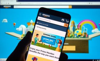 A person holds a smartphone displaying the Amazon app's homepage, with a colorful background featuring various icons and graphics.