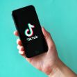 A hand holding a smartphone displaying the TikTok logo on a teal background. A hand holding a smartphone displaying the TikTok logo on a teal background.
