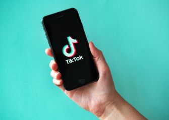 A hand holding a smartphone displaying the TikTok logo on a teal background.