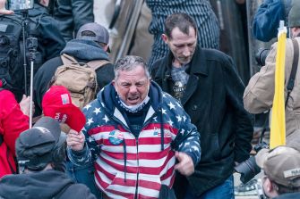 A man in a red, white, and blue hoodie, appearing emotional while surrounded by a crowd during the January 6 Capitol riot.