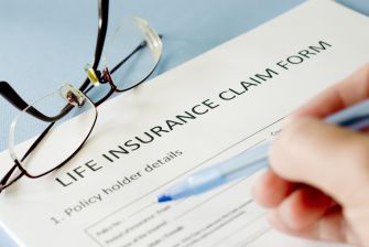 A close-up of a hand filling out a life insurance claim form, with reading glasses in the background.