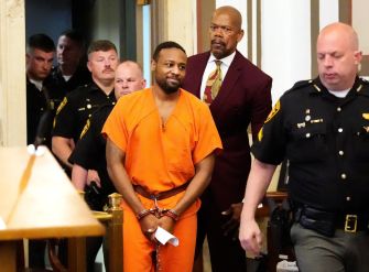 A man in handcuffs wearing an orange jumpsuit is being escorted through a courtroom by law enforcement officials.