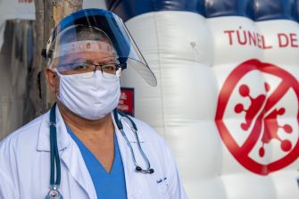 A healthcare worker wearing a mask and face shield stands in front of a COVID-19 safety tunnel.