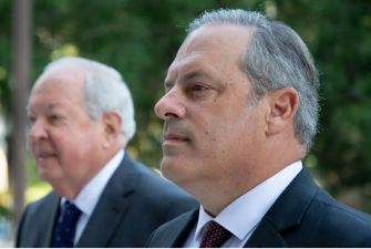 Two men standing outdoors, one in a suit with a serious expression and the other in the background.