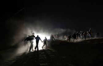 A group of individuals moving along a dusty path at night, illuminated by bright lights in the background.