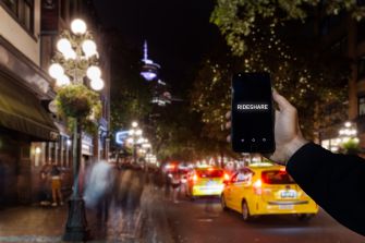 A hand holding a smartphone displaying "RIDESHARE" in a busy urban street at night, with blurred car lights and buildings in the background.