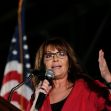 Sarah Palin speaking at an event, holding a microphone with an American flag visible in the background. Sarah Palin speaking at an event, holding a microphone with an American flag visible in the background.