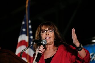 Sarah Palin speaking at an event, holding a microphone with an American flag visible in the background.