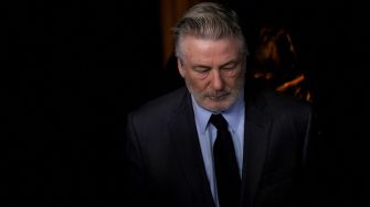 Alec Baldwin with his head down, wearing a dark suit and tie, during a serious moment.