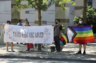 A group of protesters holding a banner reading "TRANS KIDS ARE LOVED" alongside flags supporting LGBTQ+ rights.