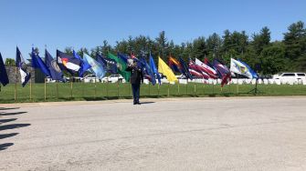 A person stands in front of a row of various state flags in a grassy area, with trees in the background.