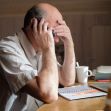 An elderly man appears distressed while on a phone call at a table, with documents and a mug nearby. An elderly man appears distressed while on a phone call at a table, with documents and a mug nearby.