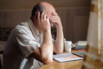 An elderly man appears distressed while on a phone call at a table, with documents and a mug nearby.