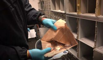 A person wearing gloves holds a box containing jewelry or valuables while inspecting empty safety deposit boxes from a private vault company.