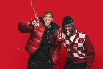 Two men posing enthusiastically while holding cans of Prime Hydration, with a splash of liquid in the air against a red background.