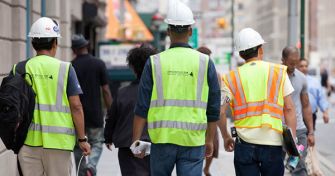 Construction workers in safety vests and hard hats walking on a busy New York City street.