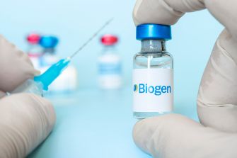 A hand holding a vial labeled with the Biogen logo, with a syringe positioned nearby, against a light blue background.