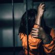 A person in an orange prison jumpsuit sitting behind bars, covering their face with their arms. A person in an orange prison jumpsuit sitting behind bars, covering their face with their arms.