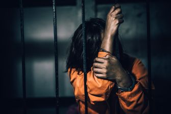 A person in an orange prison jumpsuit sitting behind bars, covering their face with their arms.