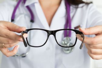 A person in a white coat holding a pair of black glasses, possibly indicating a medical or vision-related context.