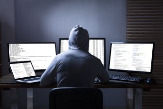 A person in a hoodie working on multiple computers, seemingly engaged in hacking activities.