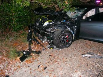 Image of a severely damaged Tesla Model S involved in a crash, with debris scattered around and visible signs of impact.