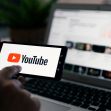 A person holding a smartphone displaying the YouTube logo, with a laptop showing YouTube's website in the background. A person holding a smartphone displaying the YouTube logo, with a laptop showing YouTube's website in the background.
