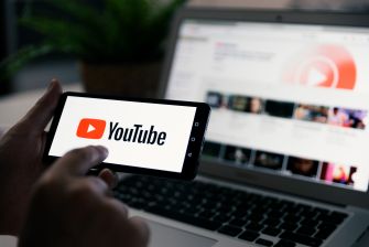 A person holding a smartphone displaying the YouTube logo, with a laptop showing YouTube's website in the background.