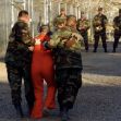 Two military personnel escorting a detainee in an orange jumpsuit at Guantanamo Bay, with other personnel observing in the background. Two military personnel escorting a detainee in an orange jumpsuit at Guantanamo Bay, with other personnel observing in the background.