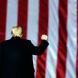 A man with blonde hair raises his fist in front of a large American flag. A man with blonde hair raises his fist in front of a large American flag.