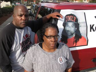 A man and woman stand in front of a vehicle displaying a poster of Kendrick Johnson, expressing their grief and determination to seek answers about his death.