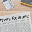 A newspaper with the headline "Press Release" on a wooden desk alongside a calculator, notebook, plants, and a cup of coffee. A newspaper with the headline "Press Release" on a wooden desk alongside a calculator, notebook, plants, and a cup of coffee.