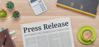 A newspaper with the headline "Press Release" on a wooden desk alongside a calculator, notebook, plants, and a cup of coffee.