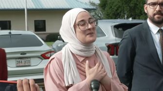 A woman wearing a hijab and glasses speaks passionately, expressing gratitude and emotion after her release from detention, with two men in formal attire standing beside her.