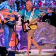 Jimmy Buffett performing on stage with a guitar, wearing a colorful shirt and shorts, in front of a band setup. Jimmy Buffett performing on stage with a guitar, wearing a colorful shirt and shorts, in front of a band setup.