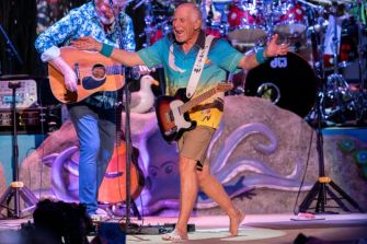 Jimmy Buffett performing on stage with a guitar, wearing a colorful shirt and shorts, in front of a band setup.