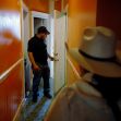 A man stands at an open door while a woman in a wide-brimmed hat approaches him in a hallway with orange walls. A man stands at an open door while a woman in a wide-brimmed hat approaches him in a hallway with orange walls.