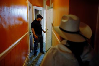 A man stands at an open door while a woman in a wide-brimmed hat approaches him in a hallway with orange walls.
