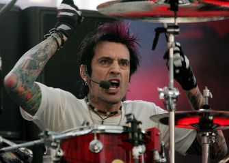 Tommy Lee performing on stage, playing the drums with intense expression and colorful tattoos visible.