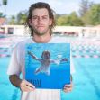 A man holding a replica of Nirvana's "Nevermind" album cover, featuring a baby swimming underwater, with a swimming pool in the background. A man holding a replica of Nirvana's "Nevermind" album cover, featuring a baby swimming underwater, with a swimming pool in the background.