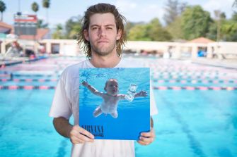 A man holding a replica of Nirvana's "Nevermind" album cover, featuring a baby swimming underwater, with a swimming pool in the background.