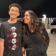 A mother and her son posing for a photo outdoors at night. The son is wearing a t-shirt with motivational text, and they are smiling at each other. A mother and her son posing for a photo outdoors at night. The son is wearing a t-shirt with motivational text, and they are smiling at each other.
