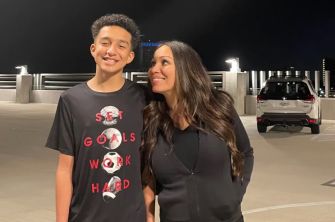 A mother and her son posing for a photo outdoors at night. The son is wearing a t-shirt with motivational text, and they are smiling at each other.