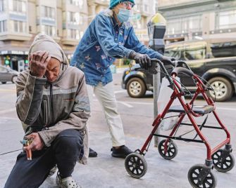 A homeless man sits on the street with his head in his hands, while a woman walking with a walker observes nearby, highlighting the struggles of the Tenderloin neighborhood in San Francisco.