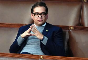 A man seated in a legislative chamber, dressed in a navy suit and glasses, looking contemplatively with hands clasped.